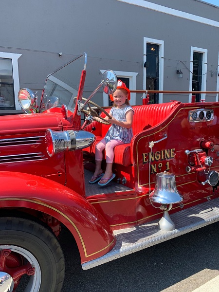 Little girl wearing a firefighter helmet sitting at the wheel of old fashioned fire truck at Geranium Festival